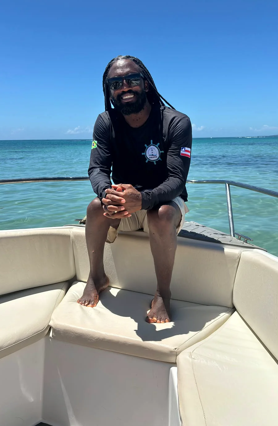 Djavan sorrindo, sentado numa lancha, óculos de sol e o mar turquesa de Morro de São Paulo ao fundo